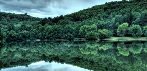 A serene lake surrounded by lush green trees, reflecting the landscape under a cloudy sky.