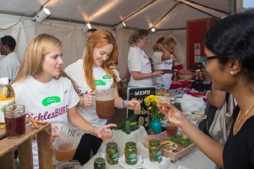 Two women serve drinks at a food festival booth, while a customer samples a beverage. Others are seen in the background.