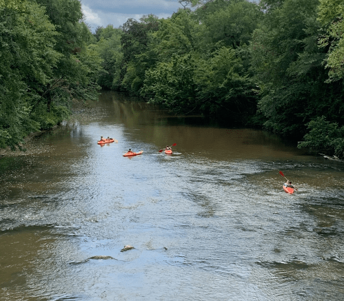 Five kayakers in orange boats paddle down a calm, tree-lined river under a cloudy sky.