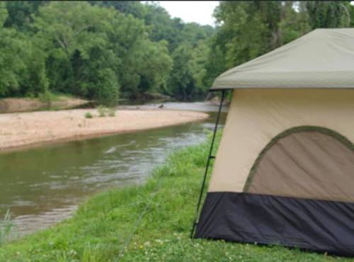 A tent set up by a calm riverbank, surrounded by lush greenery and trees.