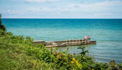 A wooden pier extends into a calm blue lake, surrounded by greenery and a clear sky. People are gathered at the end.