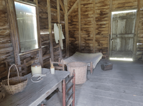 Interior of a rustic wooden room with a bed, table, basket, and a door, featuring simple furnishings and natural light.