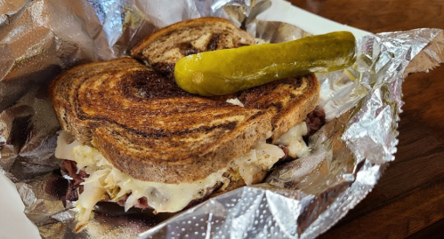 A toasted sandwich with creamy filling, wrapped in foil, topped with a pickle.