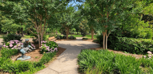 A serene park pathway surrounded by greenery and flowers, with a statue and benches inviting visitors to relax.