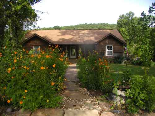 A cozy wooden house surrounded by vibrant orange flowers and a stone pathway, set against a green hillside.