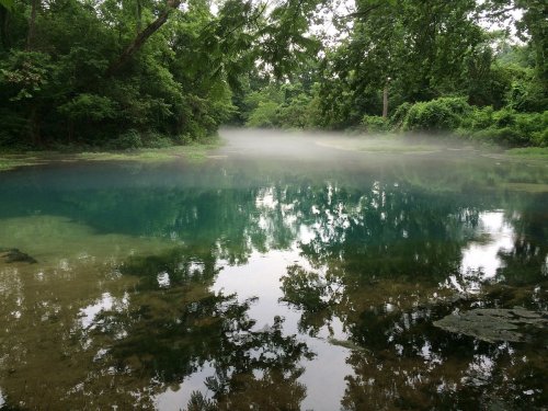 A serene lake surrounded by lush greenery, with mist rising from the water's surface and reflections of trees.