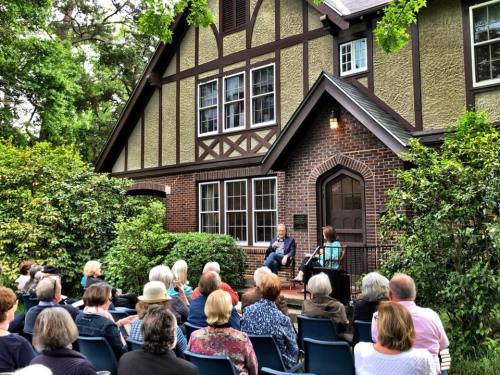 A group of people seated outdoors, listening to a speaker on a porch of a charming, Tudor-style house.