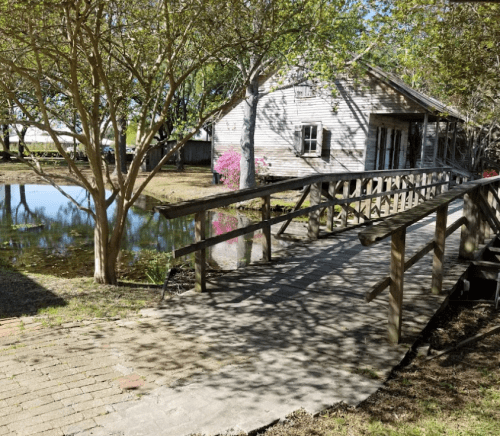 A wooden bridge leads to a rustic house by a pond, surrounded by trees and blooming flowers.