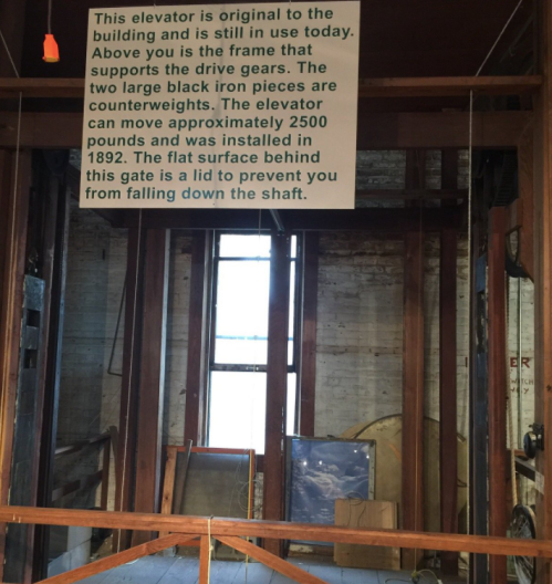 Sign explaining the elevator's history and function, with wooden beams and machinery visible in the background.