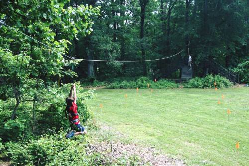 A child swings on a zip line in a wooded area, while another person waits on a platform nearby.
