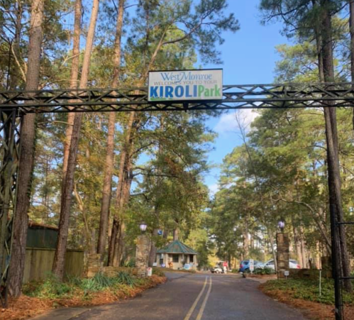 Entrance to Kiroli Park in West Monroe, surrounded by tall trees and a clear blue sky.