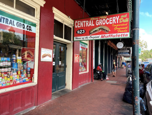 Exterior of Central Grocery with a sign featuring "House of the Original Muffuletta," showcasing colorful storefront and sidewalk.