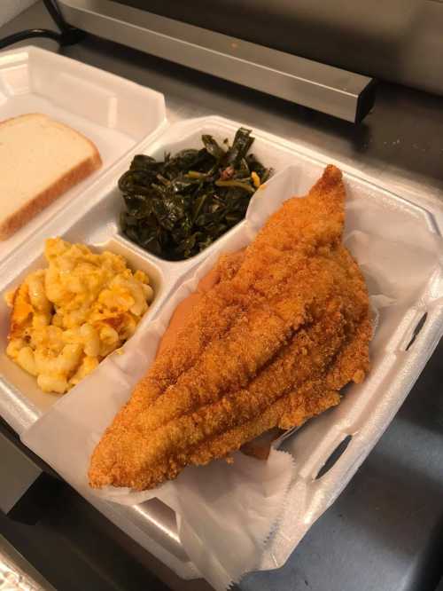 A takeout container with fried fish, collard greens, macaroni and cheese, and two slices of bread.