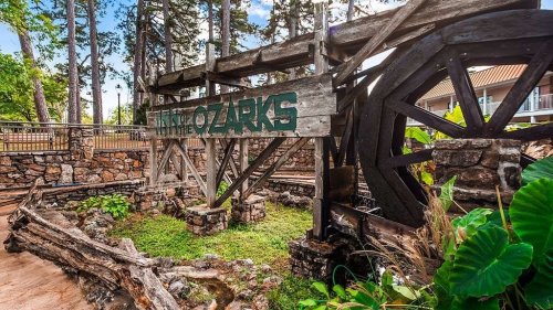 A rustic wooden sign reading "Ozarks" near a water wheel, surrounded by greenery and stone structures.