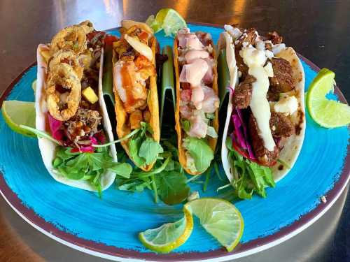 A colorful platter of four tacos with various fillings, garnished with lime wedges and fresh greens on a blue plate.