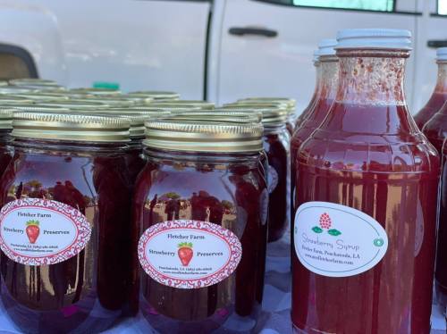 Jars of strawberry preserves and a bottle of strawberry syrup displayed at a farm stand.