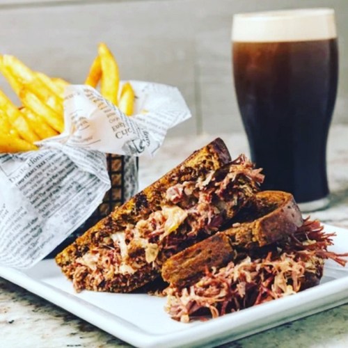 A plate with a pulled pork sandwich on toasted bread, served with fries and a glass of dark beer in the background.