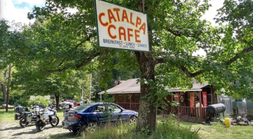 A sign for Catalpa Café hangs from a tree, with a car and motorcycles parked outside a rustic building in a wooded area.