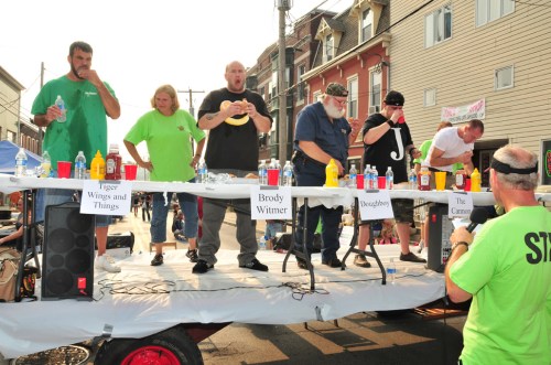 A competitive eating contest with six participants on a stage, surrounded by food and drinks, in a lively outdoor setting.