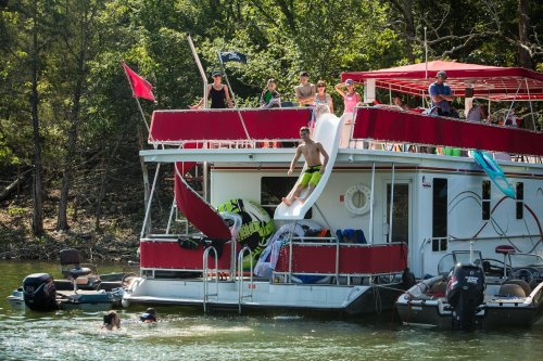 A houseboat with people on the deck and a water slide, while others swim nearby in a lake surrounded by trees.