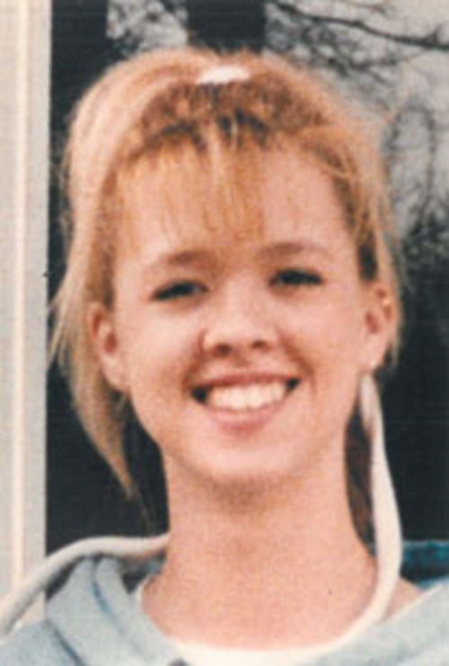 A young woman with blonde hair and bangs smiles warmly at the camera, wearing a light blue hoodie.