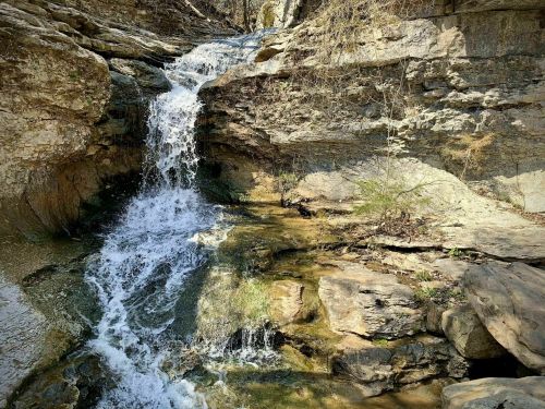 A small waterfall cascades over rocky terrain, surrounded by dry vegetation and sunlight filtering through the trees.