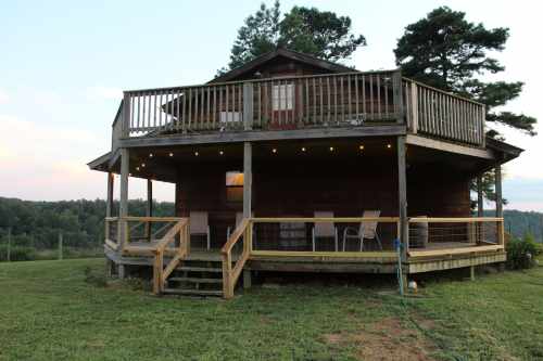 A wooden cabin with a wraparound porch, surrounded by greenery and trees, under a clear sky.