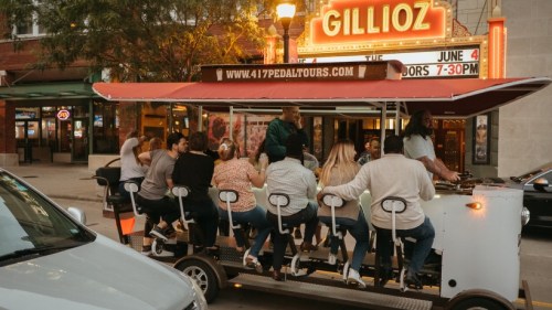 A group of people enjoying a pedal-powered tour vehicle outside a theater, with a marquee displaying an event date.