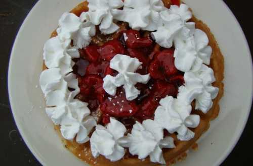 A round dessert topped with whipped cream and strawberries in a sweet sauce on a white plate.