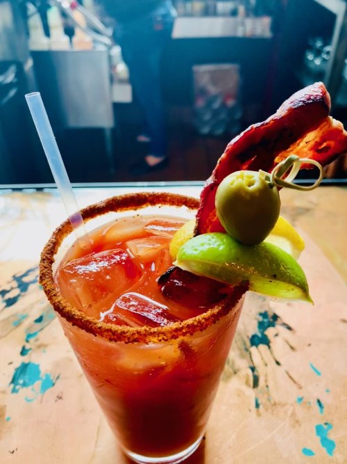 A vibrant cocktail with a bacon strip, lime, and olive garnish, served in a glass with a salted rim.