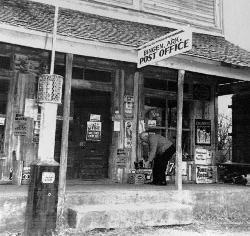 A man crouches by the entrance of a vintage post office in Bingen, Arkansas, surrounded by old signs and advertisements.