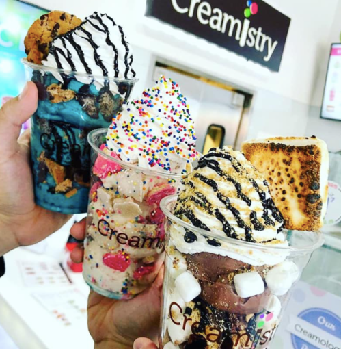 Three colorful ice cream cups topped with whipped cream, sprinkles, and cookies, held up in front of a Creamistry sign.