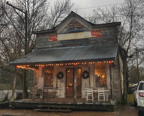 A rustic, weathered store with Coca-Cola signs, festive lights, and a porch, surrounded by trees on a cloudy day.