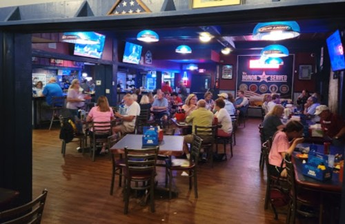 A lively restaurant interior with diners at tables, TVs showing sports, and colorful lighting.