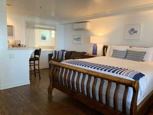 Cozy bedroom with a wooden bed, striped bedding, and a small seating area, featuring light walls and wooden floors.