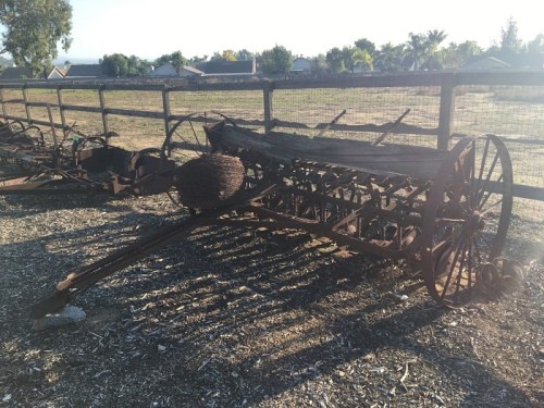 An old, rusted farming implement rests on gravel, with a wooden fence and trees in the background.