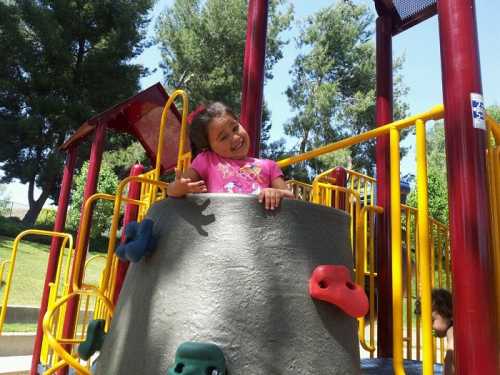 A smiling girl in a pink shirt plays on a colorful playground structure surrounded by trees.