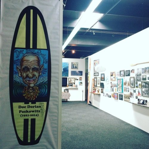 A surfboard art piece featuring Doc Dorian Paskowitz, displayed in a gallery with various framed photos on the walls.