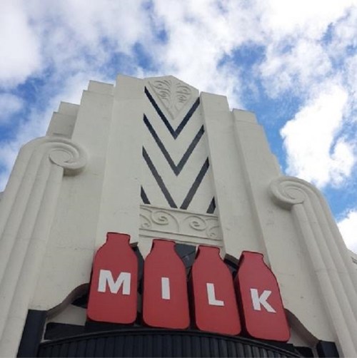 Art Deco building facade with three red milk bottle icons and the word "MILK" against a blue sky with clouds.