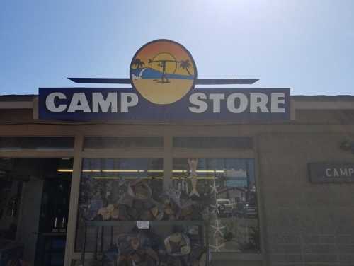 Sign for a camp store featuring a sunset with palm trees and a surfboard, with stacked wood in front.