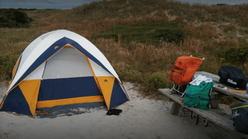 A colorful tent set up on a sandy area, with camping gear and a picnic table nearby, surrounded by grass and dunes.