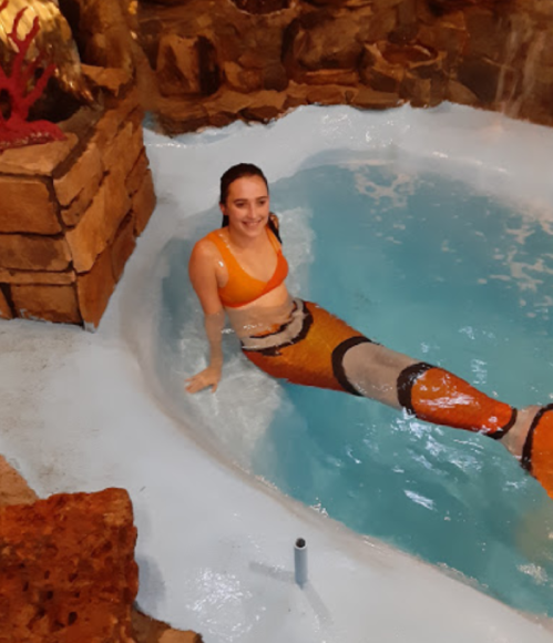 A woman in an orange bikini relaxes in a blue pool, with a colorful mermaid tail design on her legs.