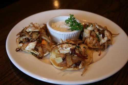 Three savory tacos topped with shredded chicken and onions, served with a small bowl of creamy dip and a garnish of parsley.