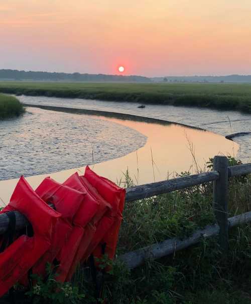 A serene sunset over a marsh, with red life jackets hanging on a fence in the foreground.