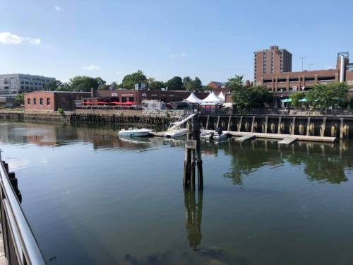 A calm river scene with docks, buildings, and tents along the shore under a clear blue sky.