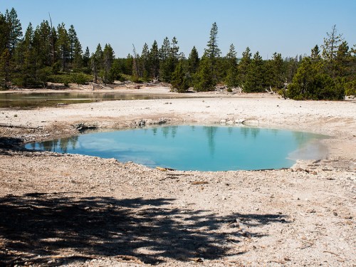 A serene blue hot spring surrounded by dry land and sparse trees under a clear blue sky.