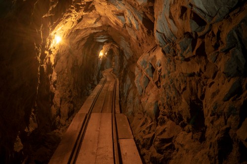 A dimly lit mine tunnel with wooden tracks running through rocky walls, illuminated by warm overhead lights.