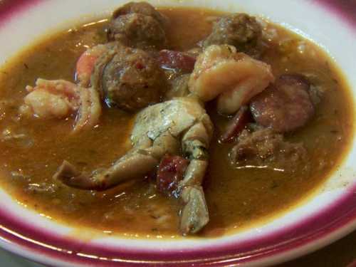 A bowl of stew featuring meatballs, shrimp, sausage, and a crab claw in a rich, brown broth.