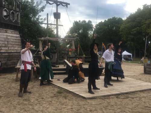 A group of performers in pirate costumes poses dramatically on a sandy stage near a wooden ship structure.