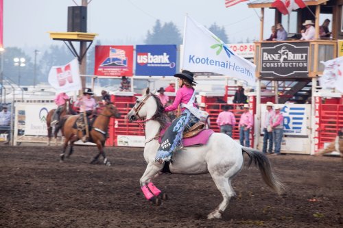 A rider in pink attire on a white horse waves a flag at a rodeo event, with spectators and other riders in the background.